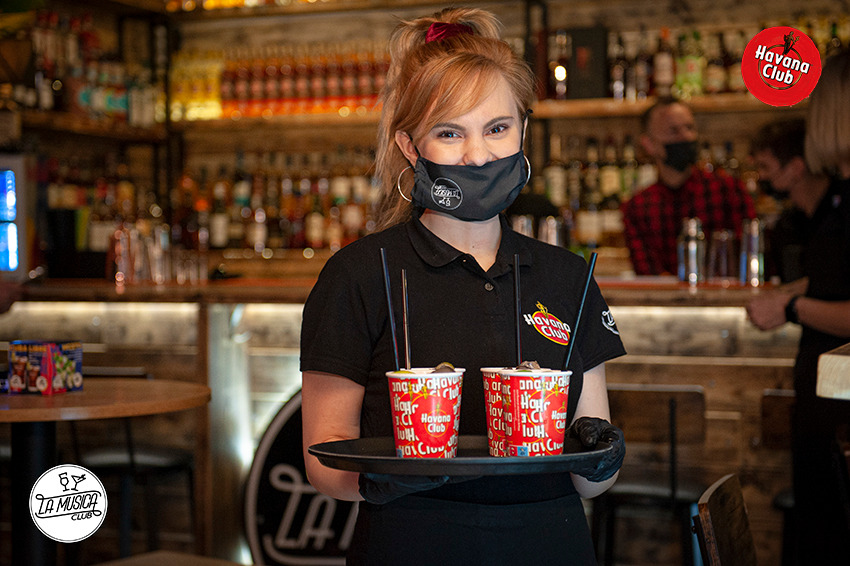 Uśmiechnięta kelnerka w czarnej masce i rękawiczkach, trzymająca tacę z dwoma drinkami Havana Club na tle baru z butelkami.